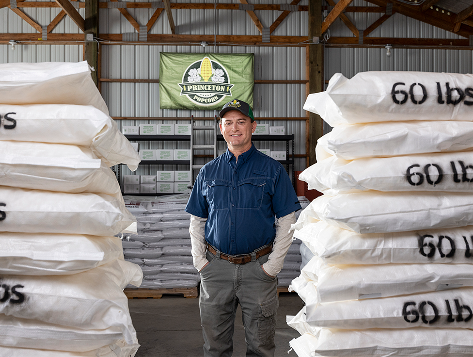 A person stands smiling in between 60-pound bags of product, with a sign in the background that says: "Princeton Popcorn."