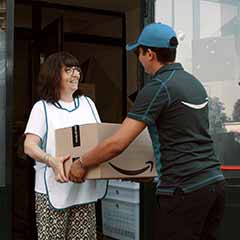 A person wearing Amazon delivery clothing hands a box to a smiling customer. 