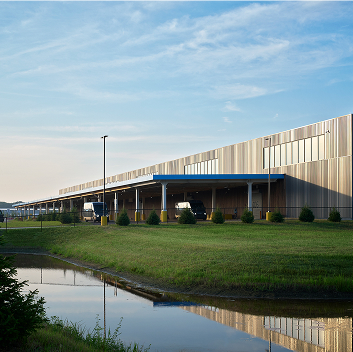 Modern industrial warehouse with two-tone facade, blue canopy over loading docks, delivery vehicles, trees, and reflecting pond in foreground.