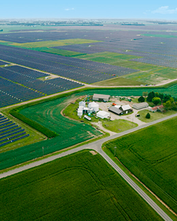Large, green fields with solar panels and building structures in the middle are shown from above. 