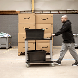 A person is shown pushing a cart past two stacks of cardboard Amazon boxes.
