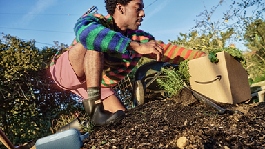 A person crouched down in a garden next to a cardboard Amazon box.\