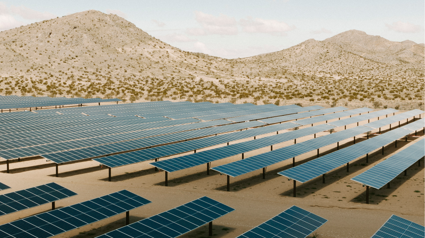 Rows of solar panels in front of rolling mountainous hills in a desert landscape.