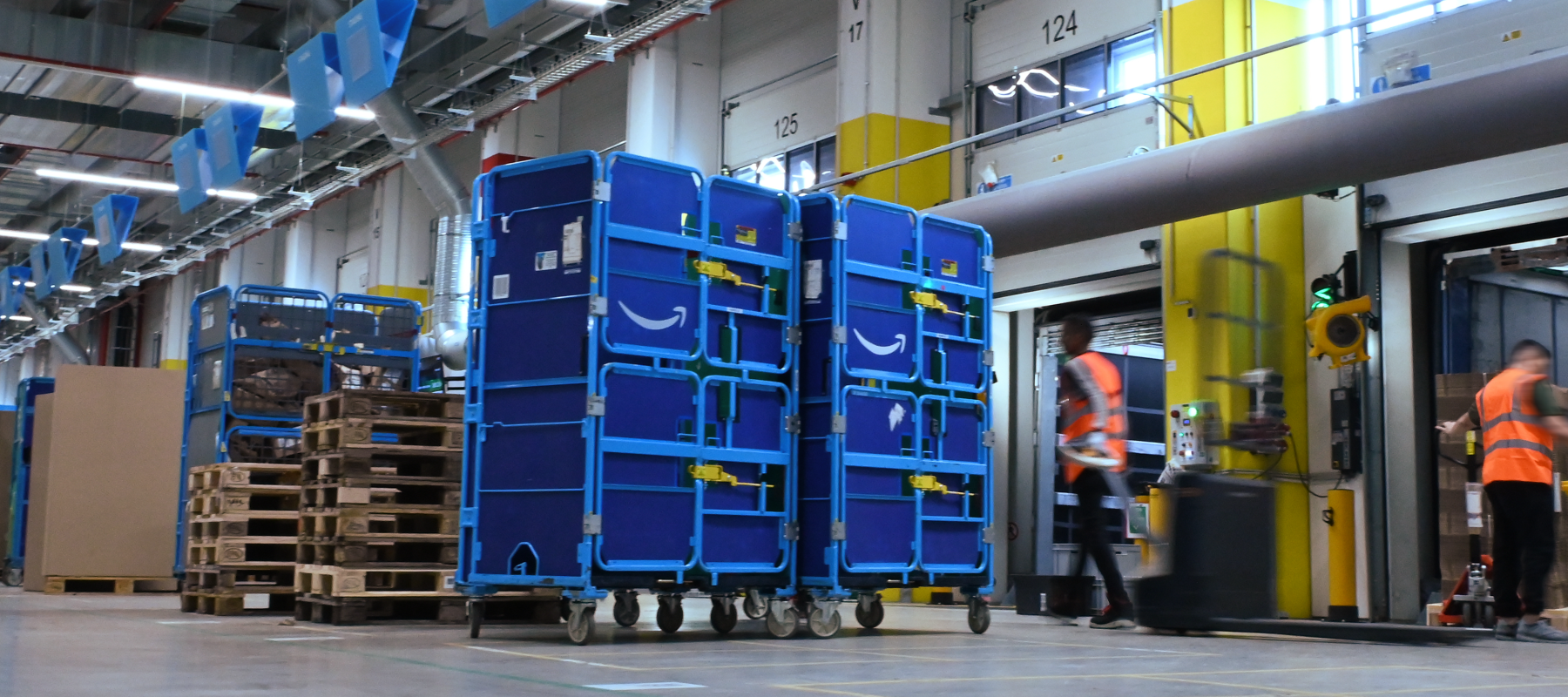 Two blue carts shown in a warehouse near a loading dock, with two people in orange vests working nearby. 