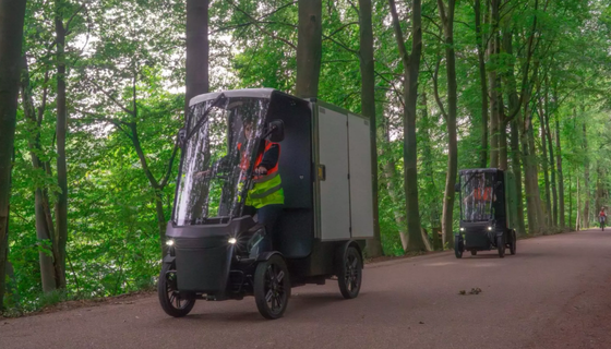 Two people drive e-bike delivery vehicles along a forest path. 