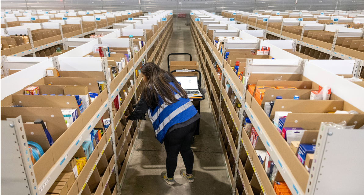 A person stands in an aisle between shelves while grabbing an item out of a bin.